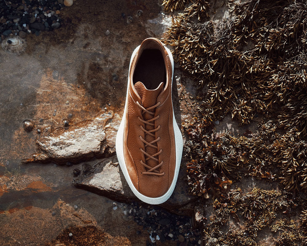 THE 1812 MIDAS SNEAKER IN TAN ON THE COASTAL ROCKS THAT INSPIRED IT, HANDMADE IN ENGLAND.